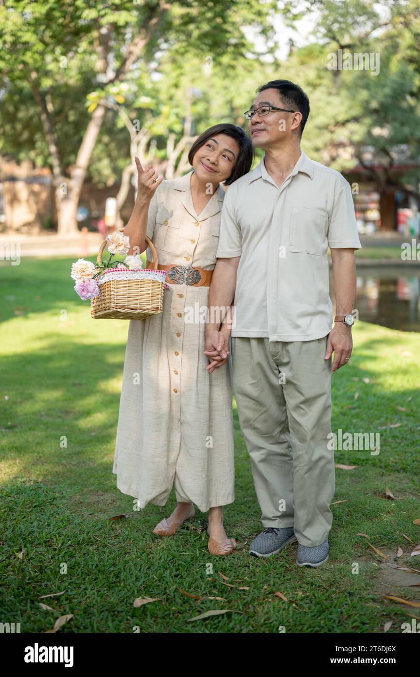 Schöne und glückliche asiatische Seniorenpaare halten Hände und genießen es, an einem hellen Tag gemeinsam durch den grünen Park zu schlendern. Glückliches Heiratsleben Stockfoto