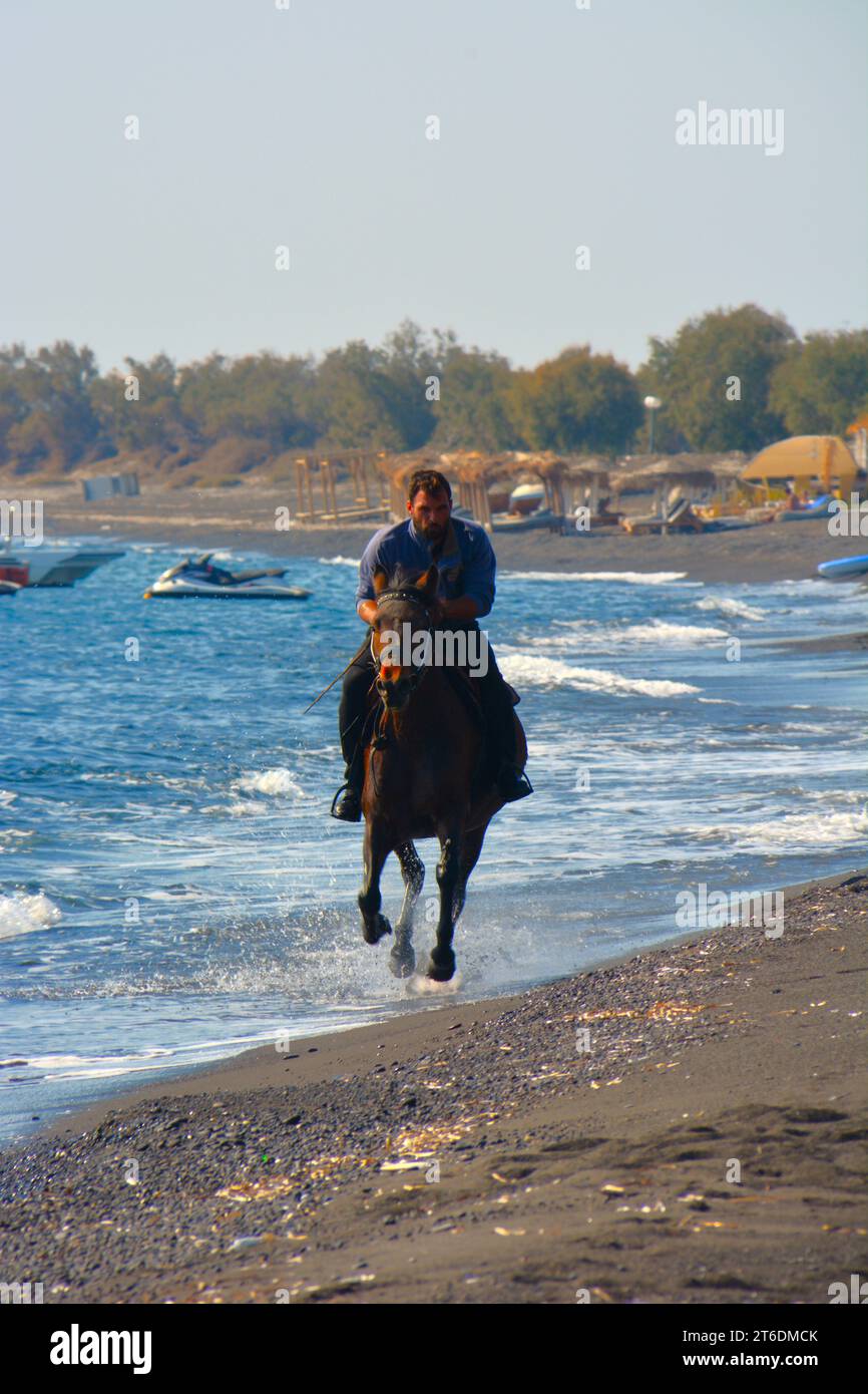 Ein Mann, der auf einem Pferd am schwarzen Sandstrand von Santorin in Griechenland reitet. Stockfoto