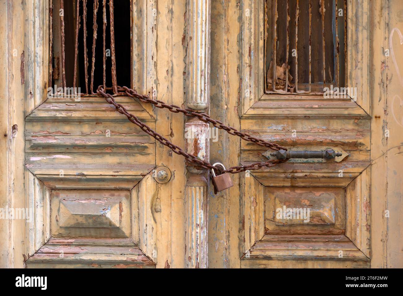 Rostige Kette und Vorhängeschloss an einem zerstörten geschlossenen Holzeingang mit kaputtem Fensterhintergrund. Verlassenes griechisches Haus mit komplizierter verschlissener Tür. Stockfoto