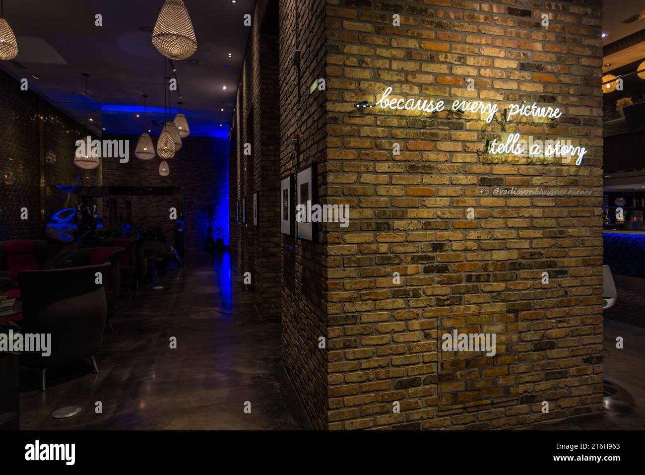 Das Neon-Schild „...weil jedes Bild eine Geschichte erzählt“ leuchtet auf einer Ziegelsäule in der Lobby des Radisson Blu Aqua Hotel in Chicago, Illinois, Vereinigte Staaten, eingerahmte Schwarz-Weiß-Fotografien der Straßenfotografin Vivian Maier säumen den Korridor dahinter. Maier arbeitete als Kindermädchen und fotografierte Chicago und New York ab den 1950er Jahren, aber ihre Arbeit wurde erst um 2014 öffentlich bekannt, nachdem Immobilienmakler John Maloof im Oktober 2009 eine Kiste mit ihren Negativen bei einer Auktion entdeckte. Das Hotel befindet sich in den unteren Etagen des Aqua Tower, der 2009 von der Architektin Jeanne Gang entworfen wurde. Stockfoto