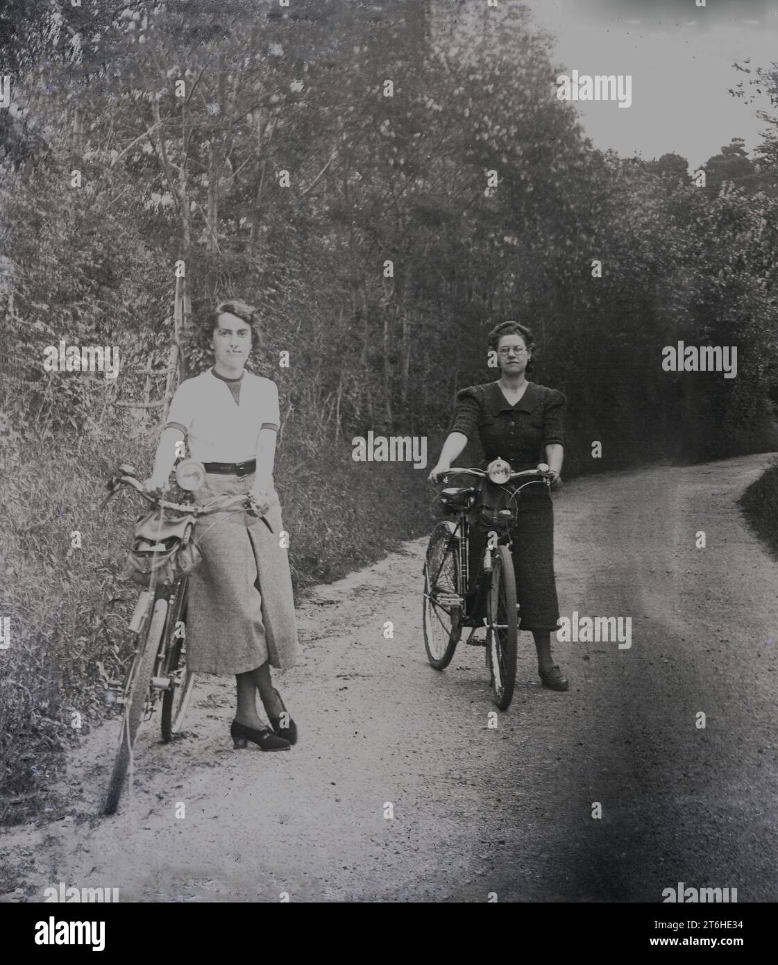 1950er Jahre, historisch, zwei junge Frauen in langen Röcken, die draußen auf einer ländlichen Gasse mit ihren Fahrrädern stehen, England, Großbritannien. Stockfoto