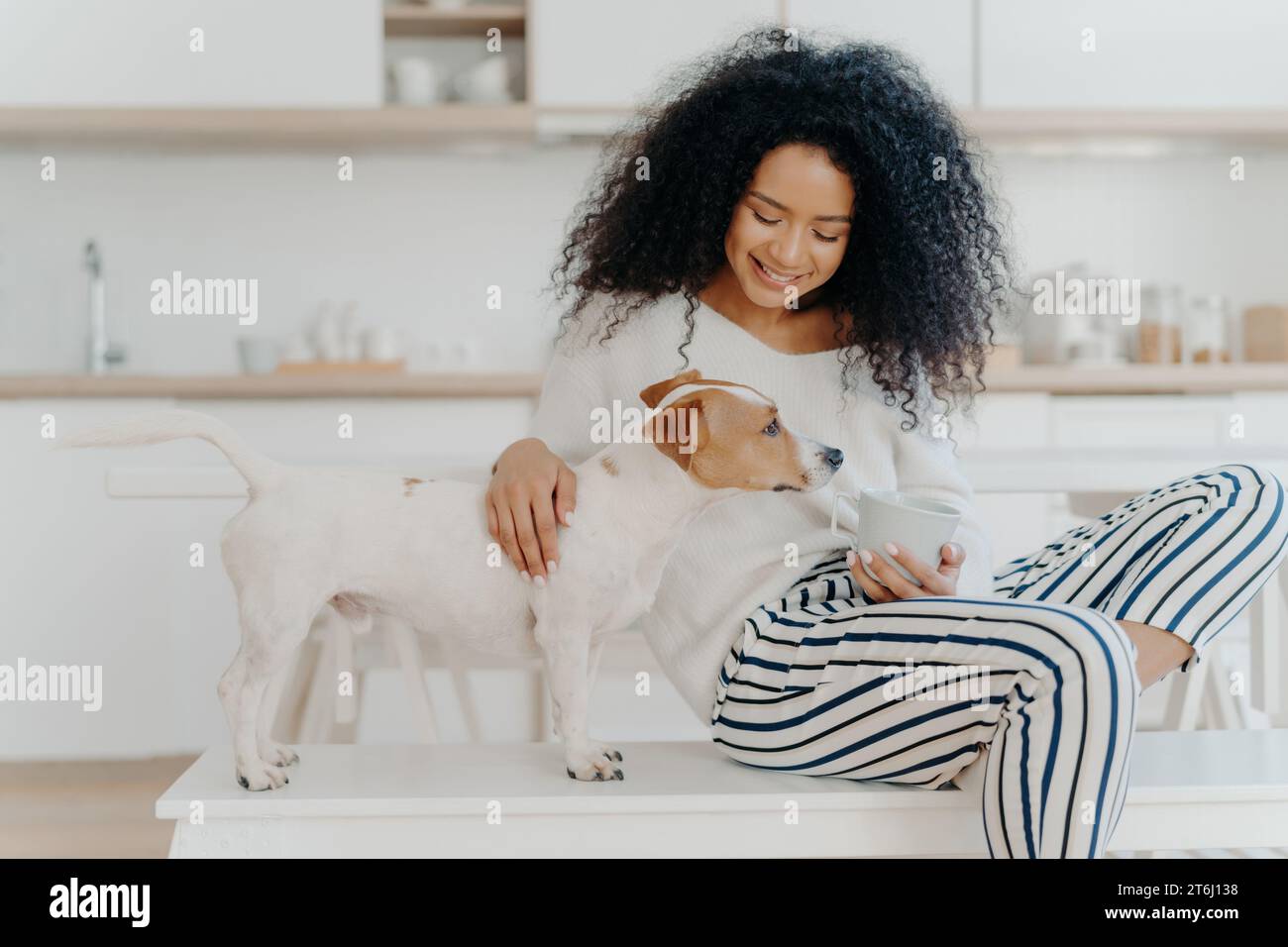 Frau mit lockigem Haar genießt einen ruhigen Moment mit ihrem Hund, eine gemütliche Szene mit einem warmen Getränk Stockfoto