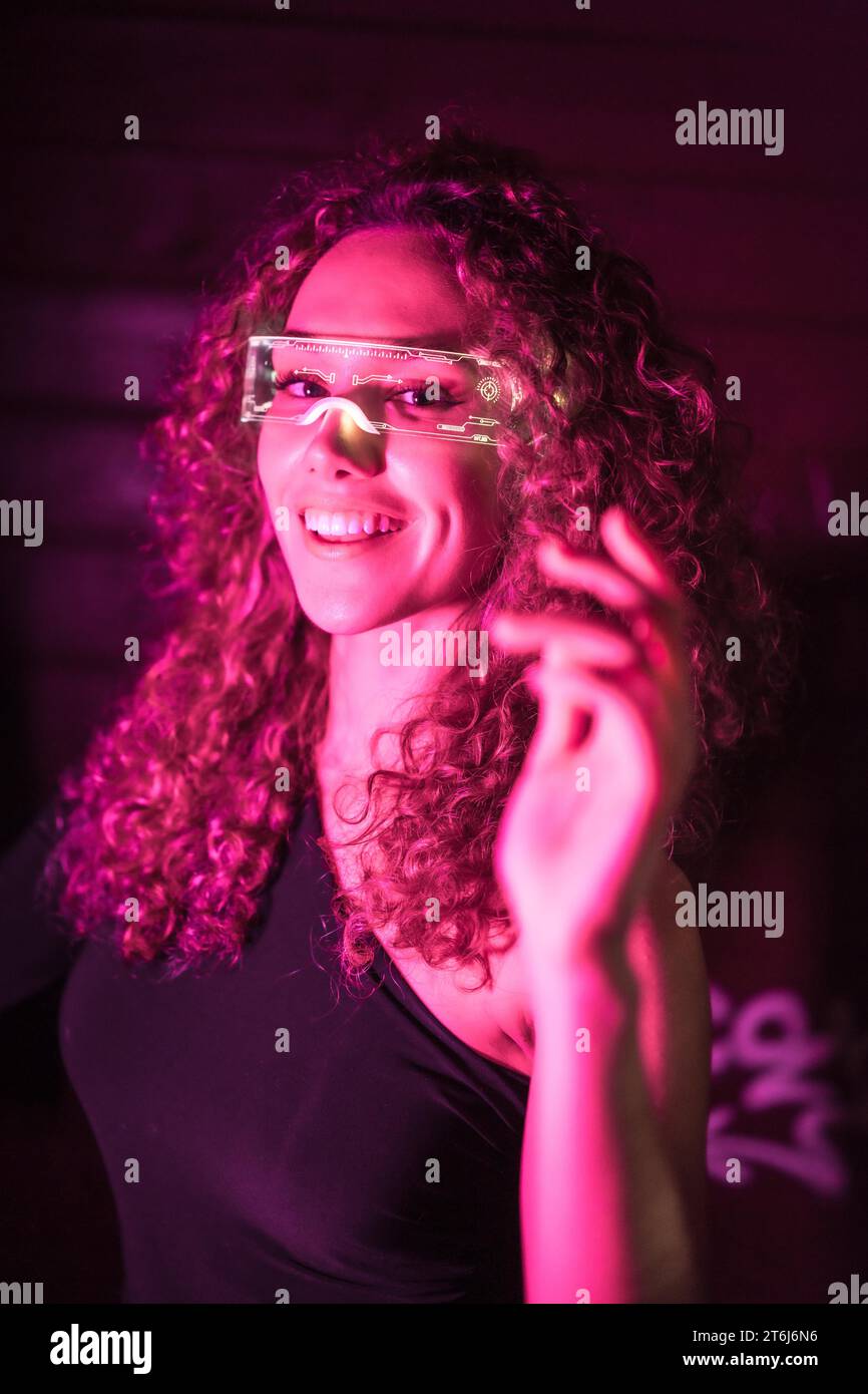 Vertikales Nahaufnahme-Porträt mit rosa Neonlichtern einer glücklichen Frau, die eine intelligente Brille trägt und in die Kamera lächelt Stockfoto