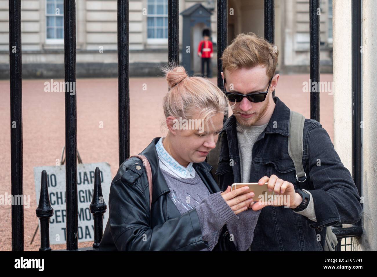 Zwei junge Leute stehen vor dem Buckingham Palace, London, während sie ein Handy mit einem Soldaten der Königsgarde im Hintergrund betrachten Stockfoto