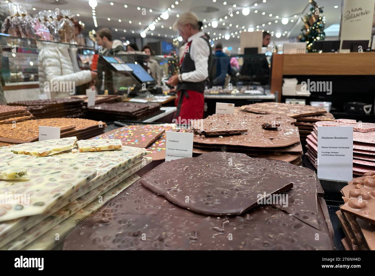 04.11.2023, Berlin, DE - Schokoladenverkauf im KaDeWe. Angebot, Auslage, Berlin, Confiserie, deutsch, Deutschland, Einzelhandel, Ernaehrung, Essen und Trinken, Europa, europaeisch, Händel, innen, Innenaufnahme, KaDeWe, Kaeufer, Kalorienbombe, Kaufhaus des Westens, Konsum, Lebensmittel, lecker, Leckerbissen, Leckerei, Menschen, Nahrung, Nascherei, Naschwerk, Personen, QF, Querformat, Schokolade, Schokoladen, Schokoladenplatten, Schokoladensorten, Schokoladenverkauf, Schokolinsen, Schweizer Milchschokolade, Sorten, Spezialitaet, suess, Suesses, Suessigkeit, Suesswaren, Verkaeuferin, Verkauf, VE Stockfoto