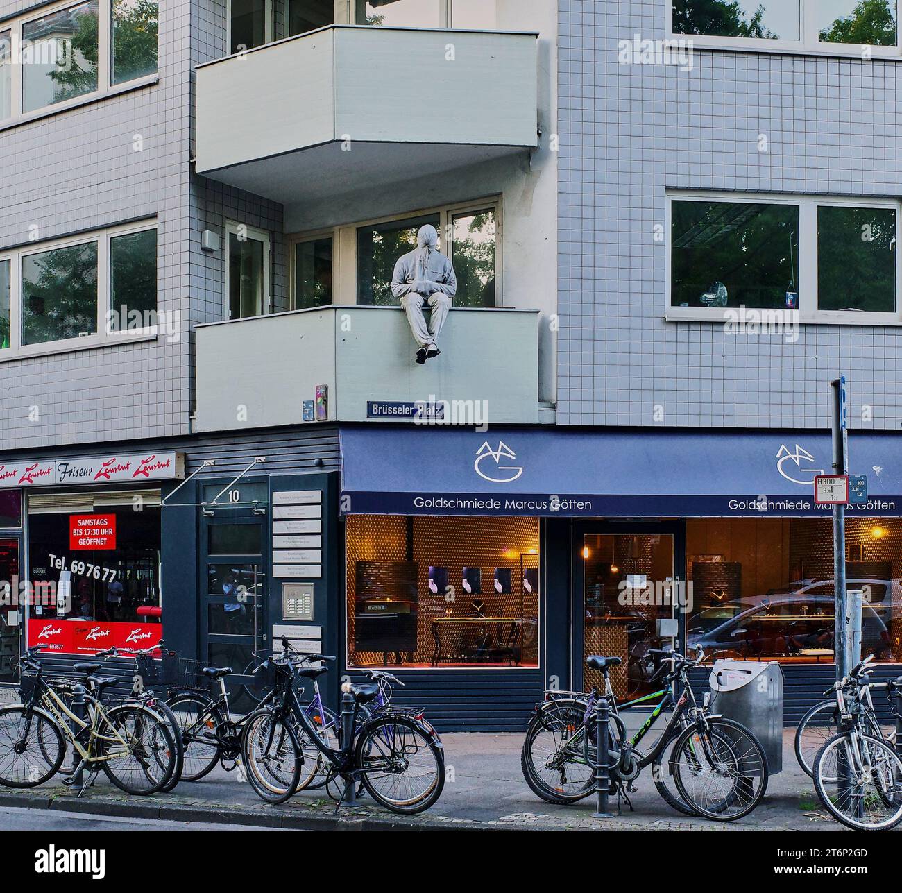 Echt oder gefälscht. Ein Mann, der wie eine Mumie eingewickelt ist, sitzt unsicher auf einem Balkon im zweiten Stock über einem Einzelhandelsgeschäft in Köln. Menschliche Kunst. Stockfoto