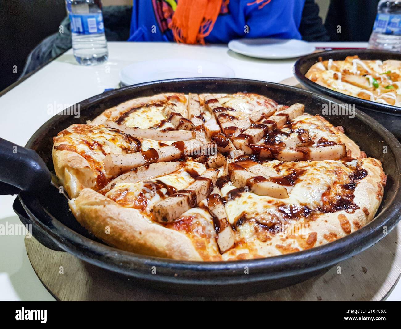 Gegrillte Hähnchenpizza auf schwarzem Teller auf weißem Tisch mit Leuten um sich herum Stockfoto