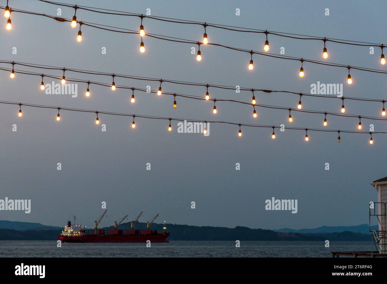 Festliche Lichter leuchten in der Abenddämmerung über Pier 11 in Astoria, Oregon, während ein großes Schiff vor der Küste im Columbia River wartet. Stockfoto