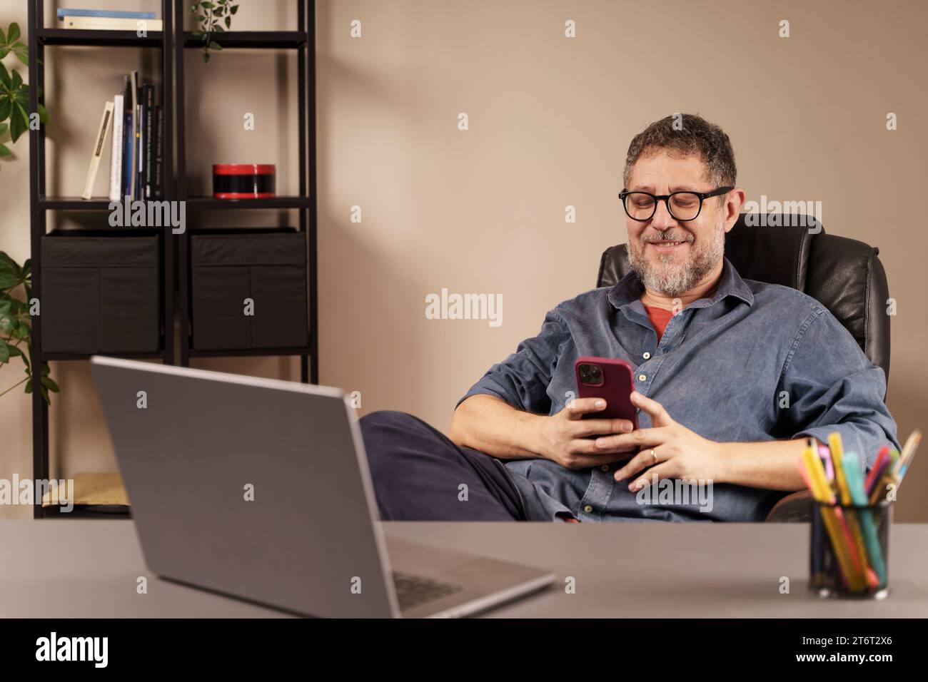 Mann mittleren Alters lächelt sein Smartphone an und arbeitet bequem von einem gut organisierten Home Office aus. Stockfoto