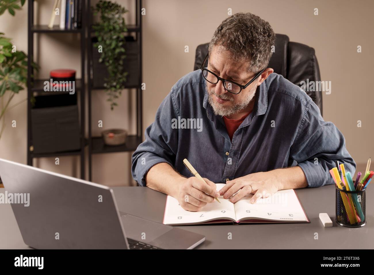 Der Mann konzentrierte sich darauf, in seinem Planer zu schreiben und Aufgaben zu organisieren, während er an seinem Laptop im Heimbüro arbeitete. Stockfoto