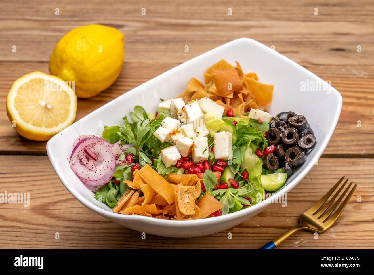 Abgebildet ist eine weiße Keramikschale mit einem bunten Salat mit einer Gabel und zwei Zitronen im Hintergrund Stockfoto