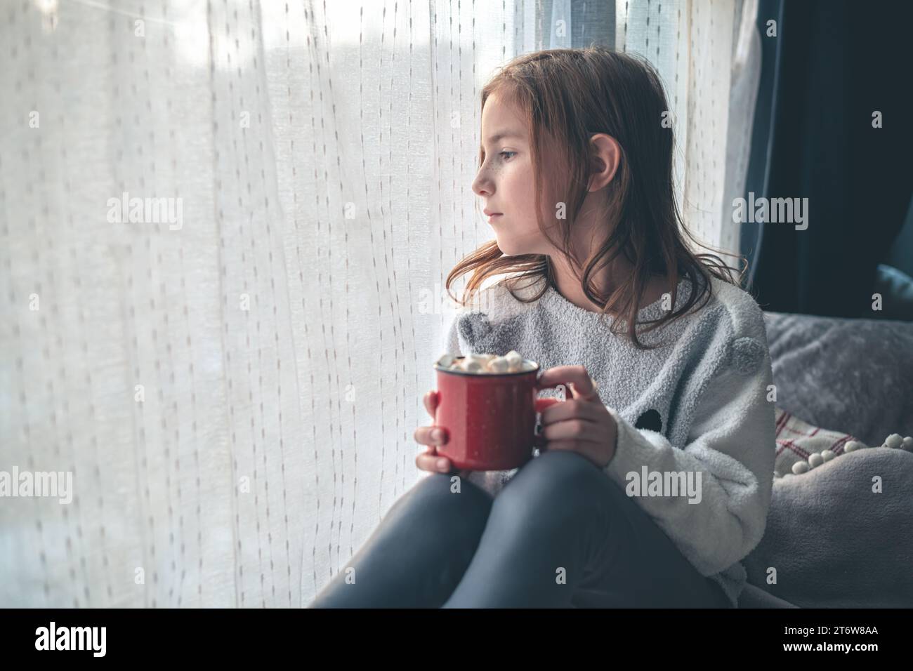 Ein süßes Mädchen auf dem Fensterbrett hält eine rote Tasse Kakao und Marshmallow und schaut aus dem Fenster. Stockfoto