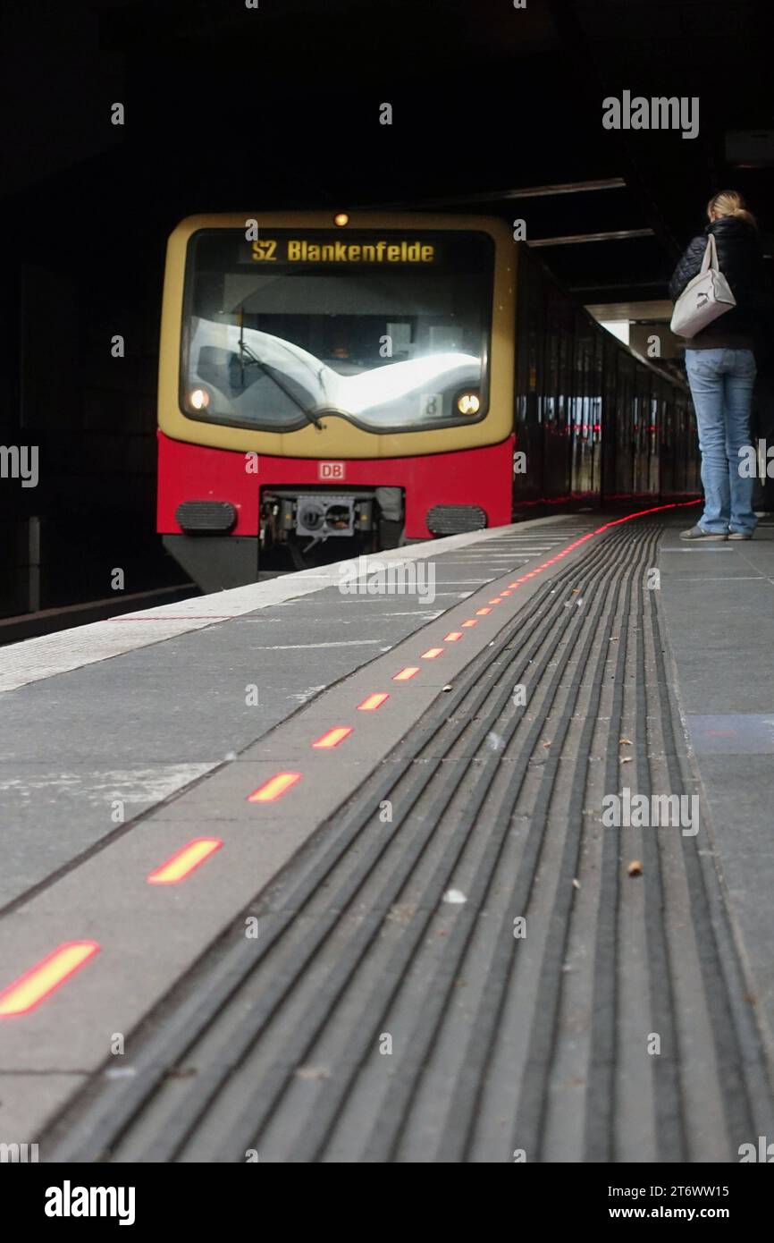 09.11.2023, Berlin, DE - Rot leuchtende Bahnsteigkante am S-Bahnhof Suedkreuz bei Einfahrt eines Zuges. Alltag, Aussen, Aussenaufnahme, Bahn, Bahnhof, Bahnstation, Bahnsteig, Bahnsteigkante, Bahnverkehr, befoerdern, Befoerderung, Berlin, deutsch, Deutschland, einfahren, Einfahrt, Europa, europaeisch, Froschperspektive, Gesellschaft, Herbst, HF, Hochformat, Jahreszeit, LED-Leitsystem, LED-Leuchten, Leitsystem, leuchtend, Lichtssteuerung, Menschen, Nahverkehr, oeffentlicher Nahverkehr, oeffentlicher Personennahverkehr, OEPNV, Personen, Personenbeförderung, Personentransport, rot, S 2, S-Bahn, Stockfoto