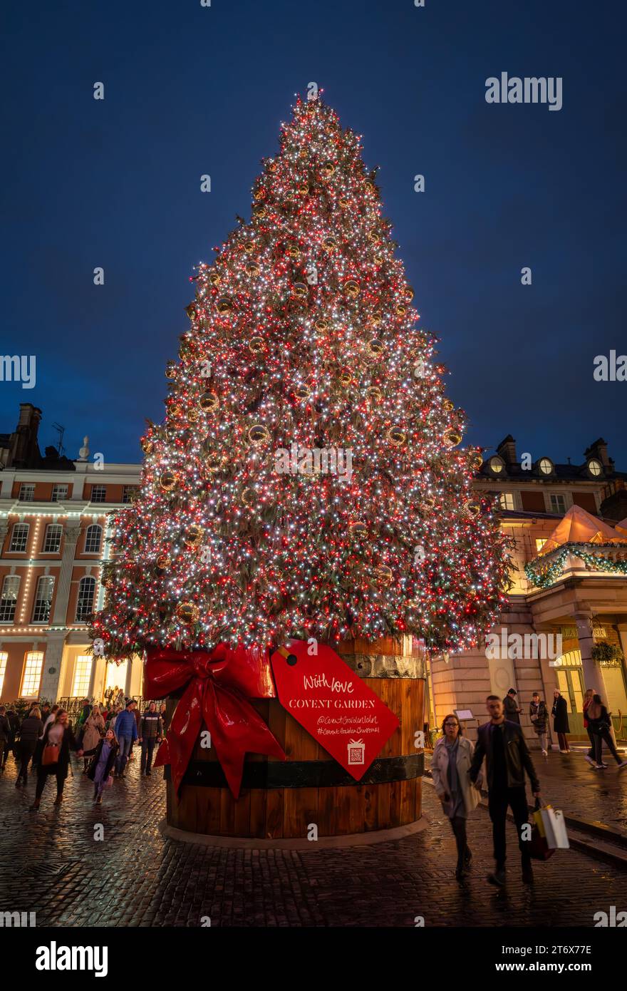 London, UK - 8. November 2023: Großer, geschmückter Weihnachtsbaum vor dem Covent Garden Market. Die Leute laufen mit Einkaufstaschen vorbei. Hochformat. Stockfoto
