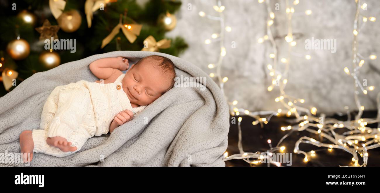 Silvester Weihnachtsfeierszene - niedlicher schlafender neugeborener Junge in einem beigefarbenen Strickanzug, der auf einer Decke liegt, mit Weihnachtsbaumspielzeugen und Lichtergirlande. Kleinkinder, Kinderzimmer Stockfoto