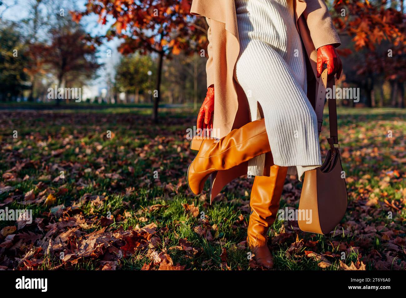 Modische Frau in stilvollem weißem Strickkleid, kniehohen Stiefeln mit Handtasche im Park. Herbstschuhe und Accessoires. Nahaufnahme der Stiefel Stockfoto