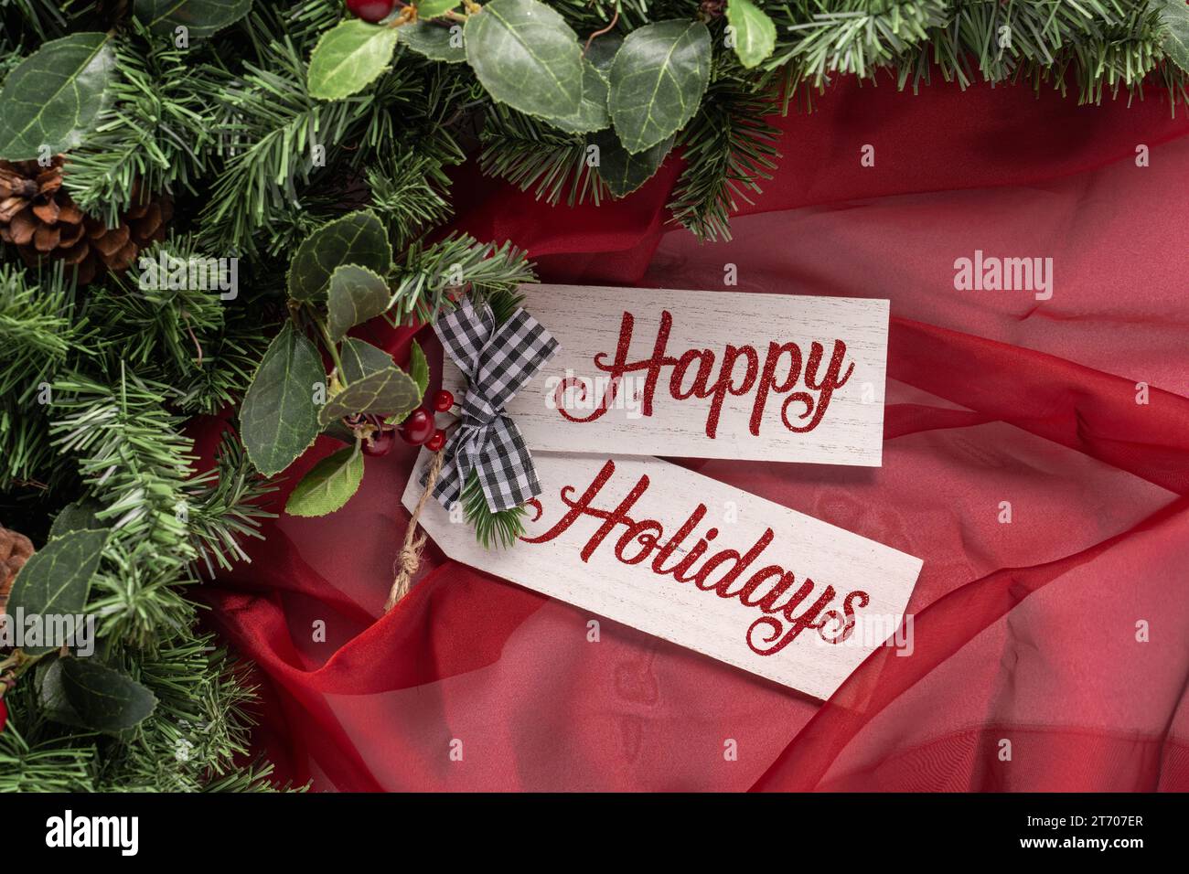 Happy Holidays Text auf Holzbrett mit transparentem rotem Stoff, Grün und einer kleinen schwarz-weißen Schleife Stockfoto