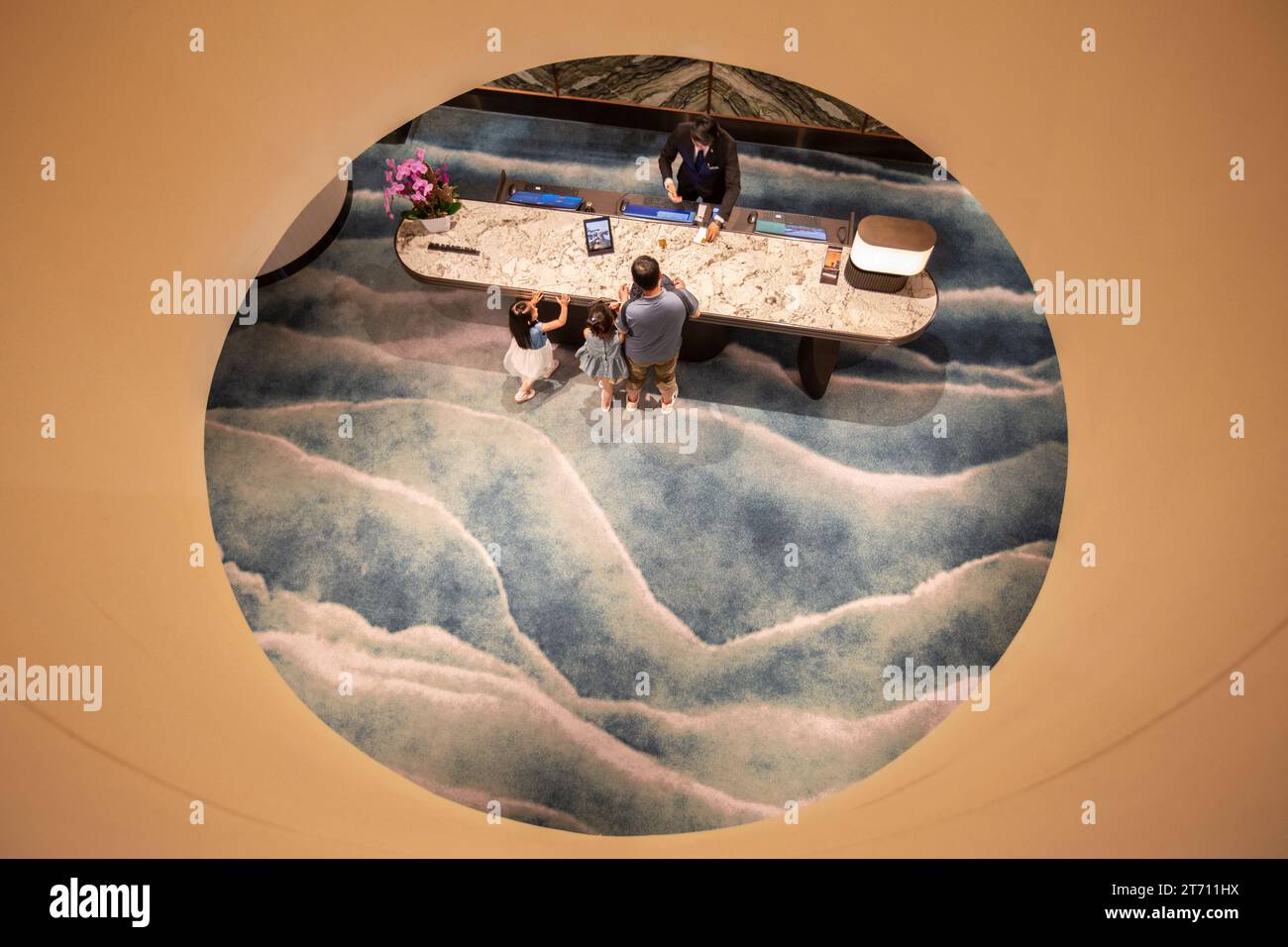 Blick von oben auf einen Mann und seine Kinder beim Einchecken in einem luxuriösen 5-Sterne-Hotel, der hellblaue Teppich zeigt die Meereswelle. Pan Pacific Hotel, Singapur. Stockfoto