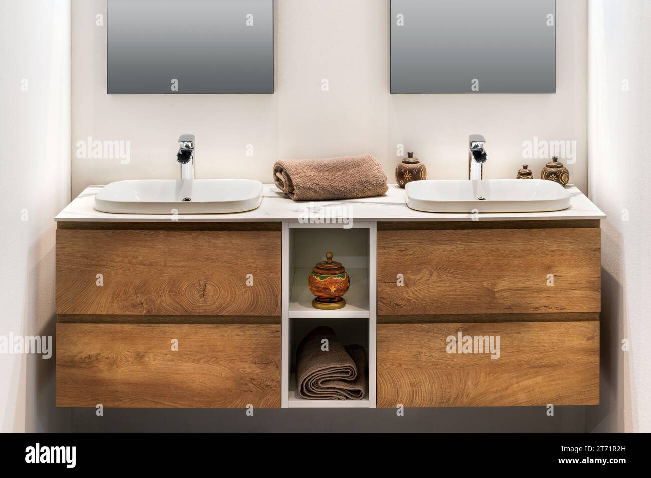 Das Innere des modernen, minimalen Badezimmers mit zwei weißen Waschbecken, Wasserhähnen, Handtuch auf Holzschrankregalen mit Räucherbrenner Vase, während mirro Stockfoto