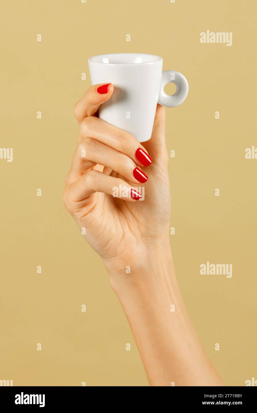 Anonyme junge schlanke weibliche Hand mit polierten Nägeln von schlanken Fingern, die eine Kaffeetasse in Licht vor braunem Hintergrund halten Stockfoto