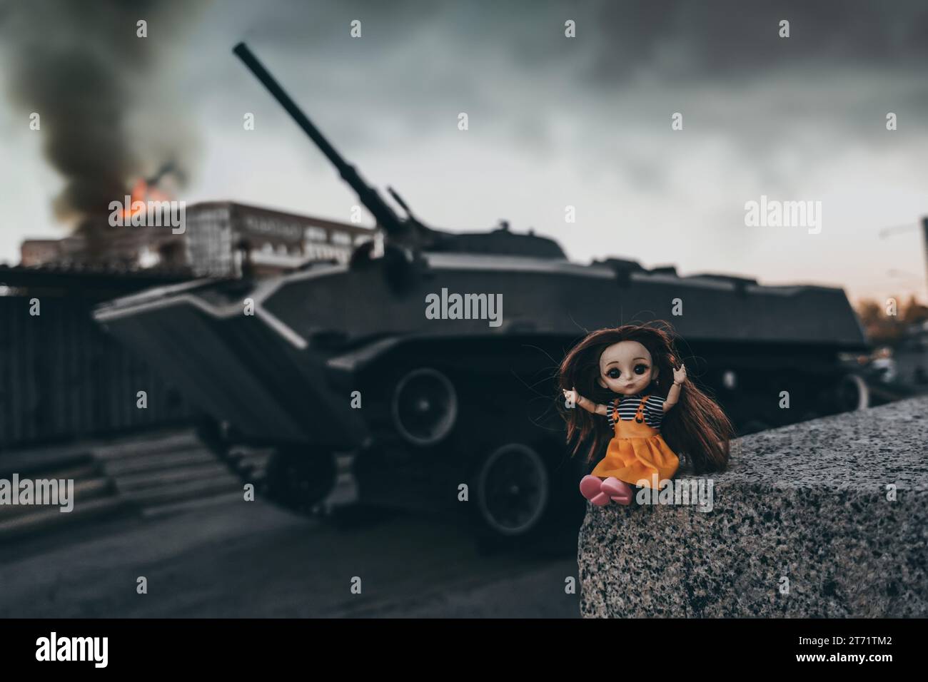 Die Kriegskrankheit zerstörte eine verängstigte Puppe vor dem Hintergrund eines Panzers und eines ausgebrannten Gebäudes. Der Begriff des Leidens von Kindern und Zivilisten Stockfoto