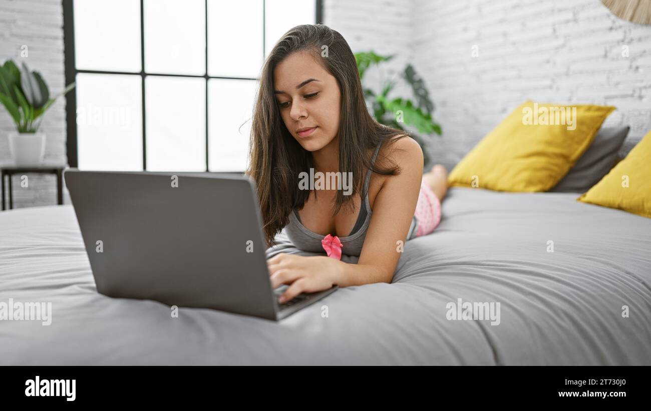 Entspannte Morgenstimmung mit einer jungen, schönen hispanischen Frau, die wach und konzentriert liegt, vertieft in ihrem Laptop auf dem gemütlichen Bett in ihrem Zimmer. Entspannte Ho Stockfoto Entspannte Morgenstimmung mit einer jungen, schönen hispanischen Frau, die wach und konzentriert liegt, vertieft in ihrem Laptop auf dem gemütlichen Bett in ihrem Zimmer. Entspannte Ho Stockfoto