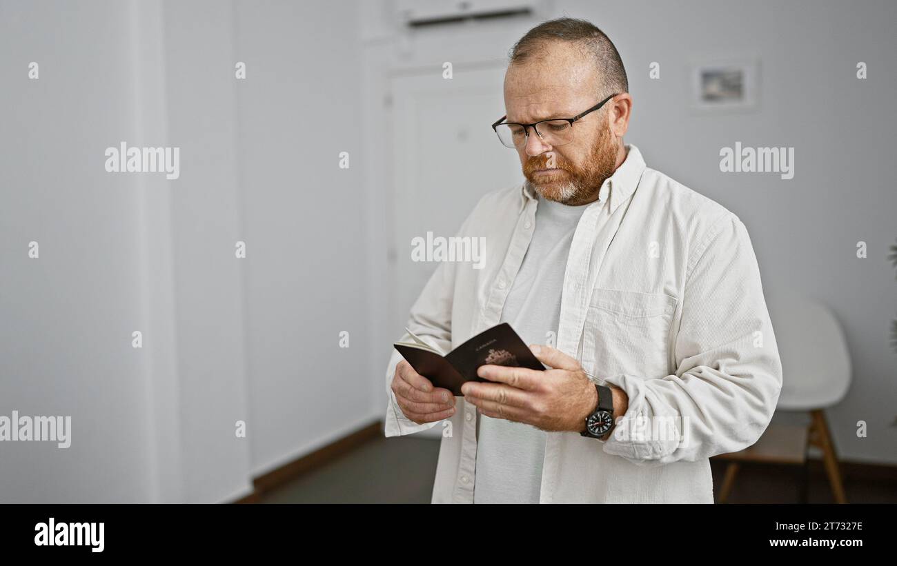 Gutaussehender kaukasier mittleren Alters, mit Bart und Brille, hält seinen Pass, in Gedanken getaucht, während er lässig im Wartezimmer steht, po Stockfoto