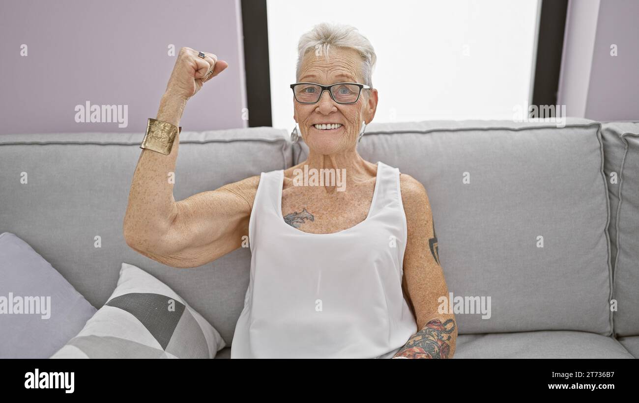 Selbstbewusste grauhaarige Seniorin, die ihren Bizeps auf dem Wohnzimmer-Sofa zu Hause fröhlich biegt, mit einem großen, fröhlichen Lächeln und einem coolen, positiven Akt Stockfoto