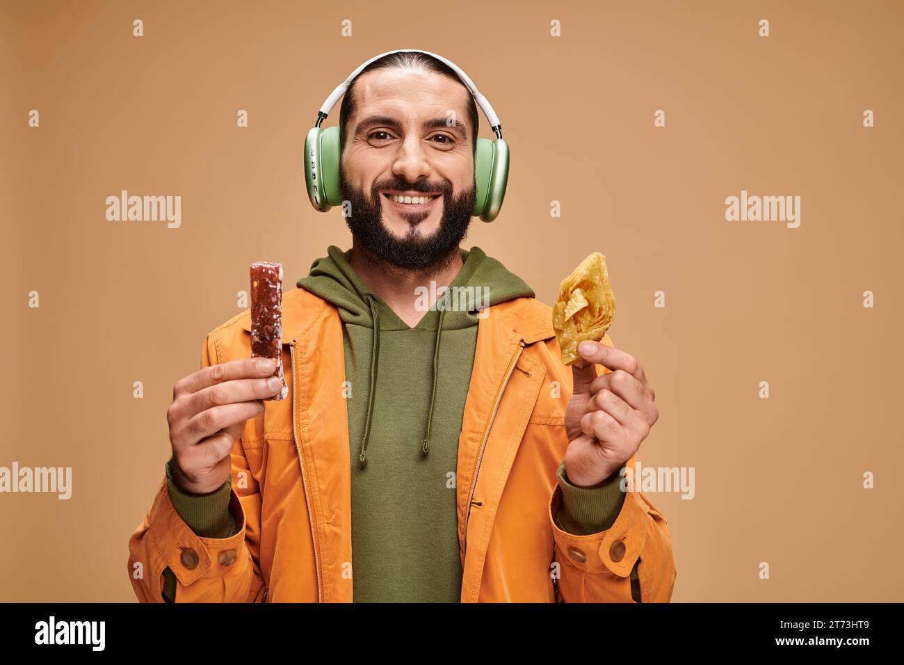 Glücklicher arabischer Mann in Kopfhörern mit zwei Desserts aus dem Nahen Osten, Honigbaklava und Kirchchchela Stockfoto
