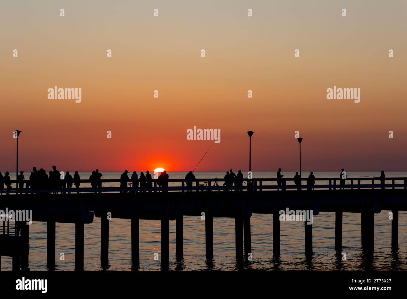 Dunkle Umrisse von Menschen, die während der untergehenden Sonne auf dem Seebrück spazieren gehen. Der Himmel leuchtete orange mit den letzten Sonnenstrahlen. Stockfoto