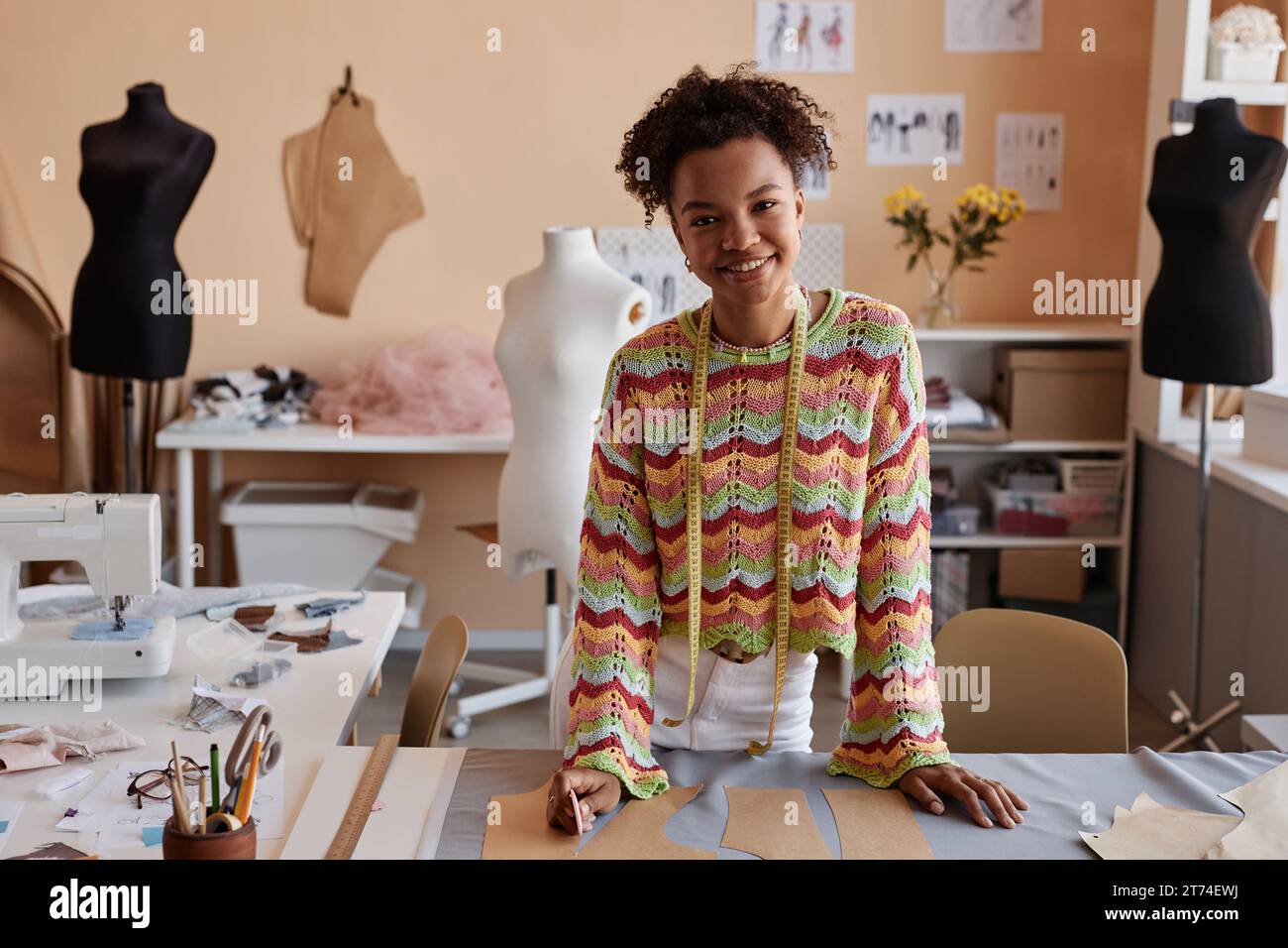 Junge lächelnde Handwerkerin in Casualwear, die am Arbeitsplatz in einem Studio für Modedesign oder Schneiderei steht und in die Kamera schaut Stockfoto