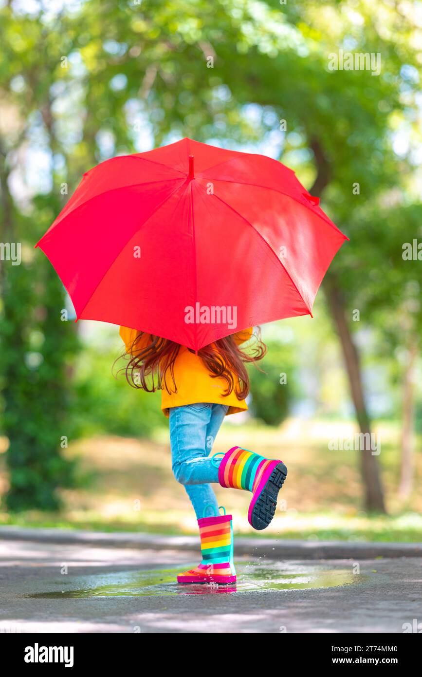 Glückliches, wunderschönes Mädchen mit Regenschirm und farbigen Gummistiefeln springt auf eine regnerische Pfütze Stockfoto