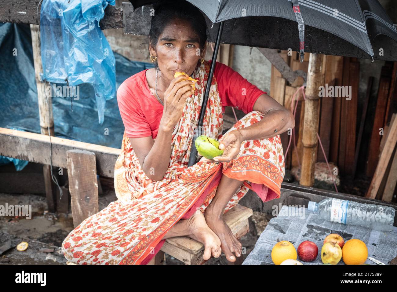 Frau isst eine Mango vor ihrem Haus, schützt sich mit einem Regenschirm vor einer Monsundusche, Tejgaon Slum Gegend, Dhaka, Bangladesch Stockfoto