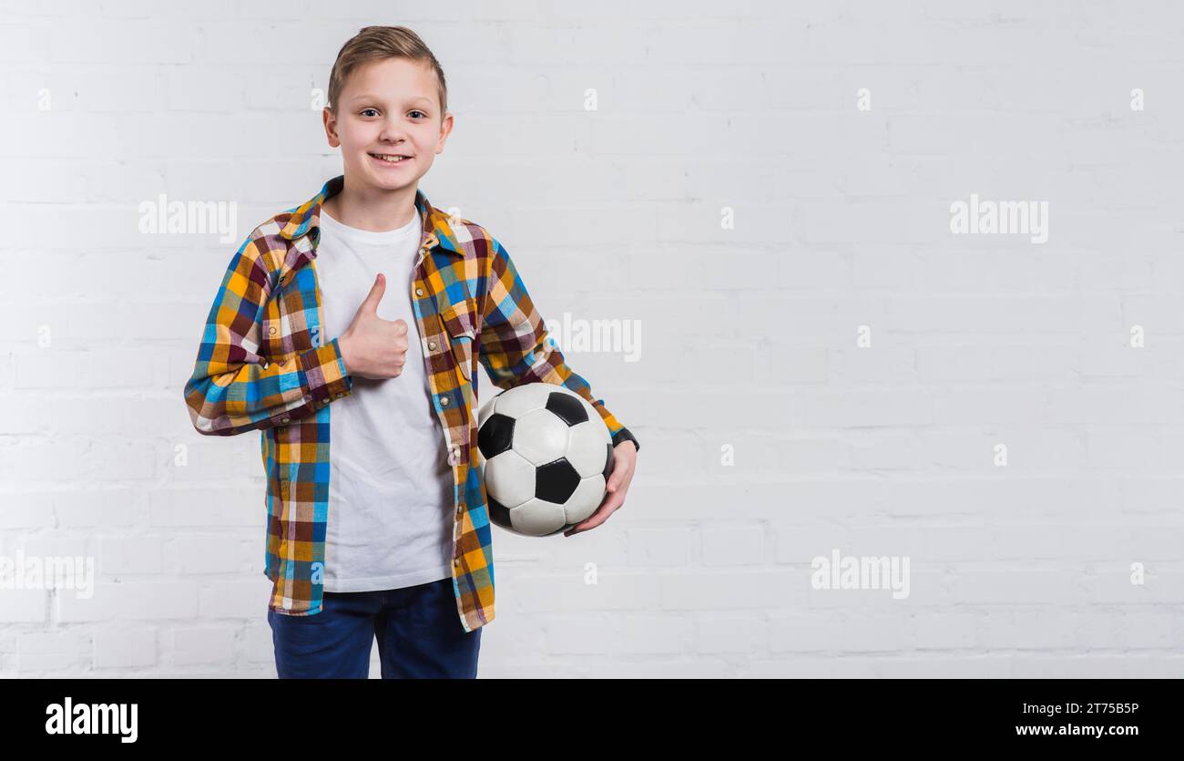 Ein lächelnder Junge, der die Fußballhand hält, zeigt ein Schild mit dem Daumen nach oben, das an einer weißen Ziegelwand steht Stockfoto