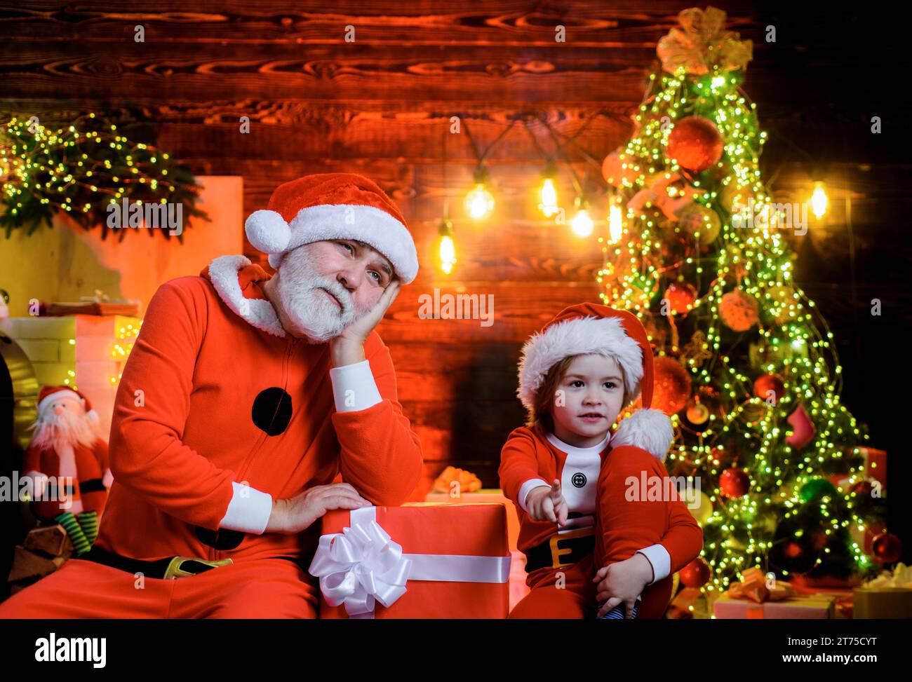 Weihnachtskind mit Weihnachtsmütze und Weihnachtsmann mit Weihnachtsgeschenk zu Hause. Weihnachtsmann und kleiner Junge mit Weihnachtsgeschenk. Junge und bärtiger Mann drin Stockfoto