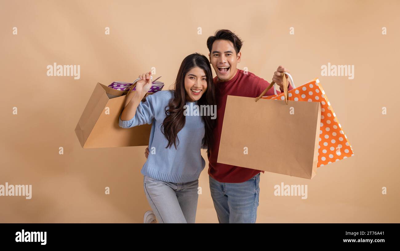 Glückliches lächelndes asiatisches Paar mit Einkaufstaschen im Pullover, das ihren Winterrabatt-Verkauf auf beigefarbenem Hintergrund genießt. Stockfoto