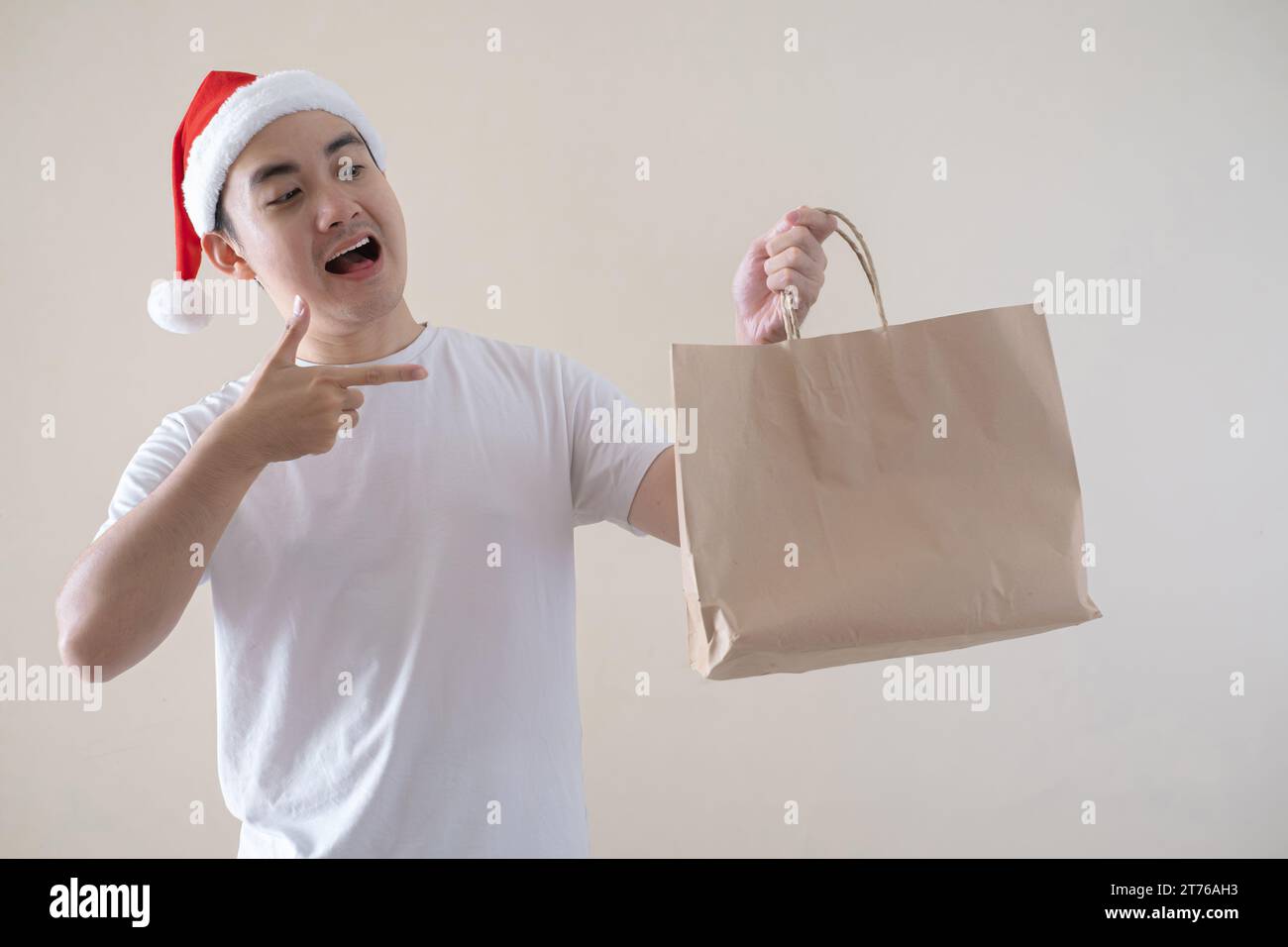 Der junge asiatische Weihnachtsmann zeigt auf die Einkaufstasche auf isoliertem beigefarbenem Hintergrund. Weihnachtskonzept. Stockfoto