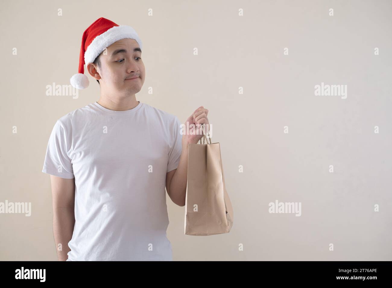 Der junge asiatische Weihnachtsmann hält sich an der Einkaufstasche auf isoliertem beigefarbenem Hintergrund. Weihnachtskonzept. Stockfoto
