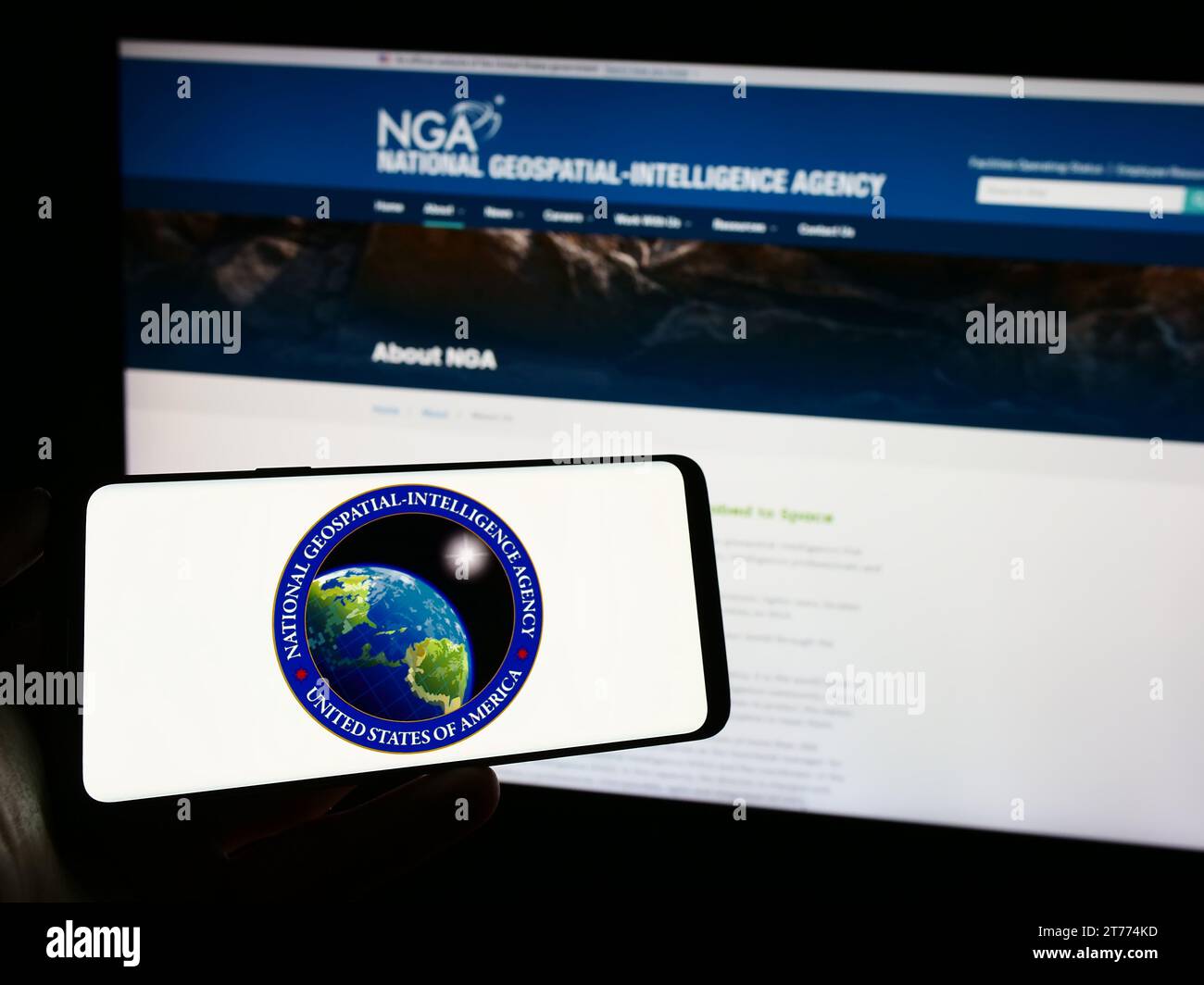 Person, die ein Mobiltelefon mit dem Siegel der US National Geospatial-Intelligence Agency (NGA) vor der Webseite hält. Schwerpunkt auf dem Display des Telefons. Stockfoto