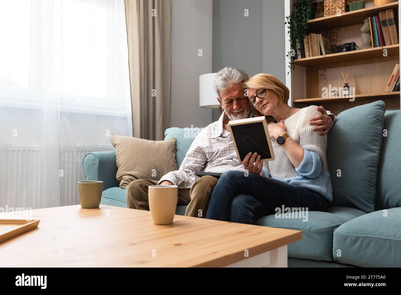 Zwei ältere Leute, ein älteres Paar, Mann und Frau, die ein Familienfoto ansehen, lächeln und umarmen, während sie zu Hause auf dem Sofa sitzen. Erinnerungen und Nostalgie für das Kind Stockfoto