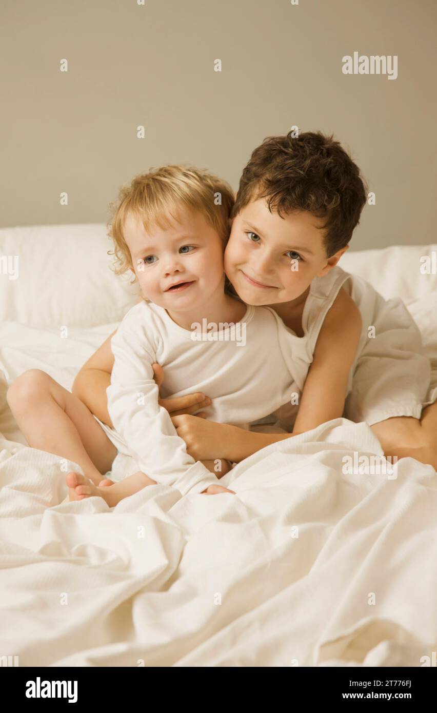 Junge und Kleinkind sitzen im Bett umarmt und lächelnd Stockfoto