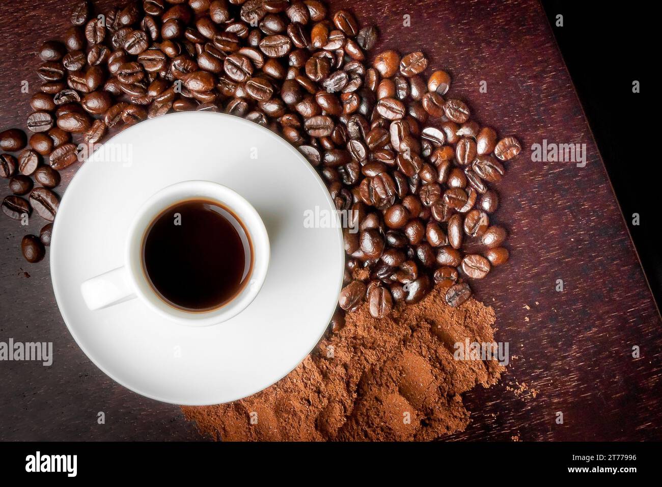 Blick von oben auf eine weiße Tasse mit Kaffee in der Nähe von Kaffeebohnen und Kaffeepulver über einem holzbraunen Tisch Stockfoto