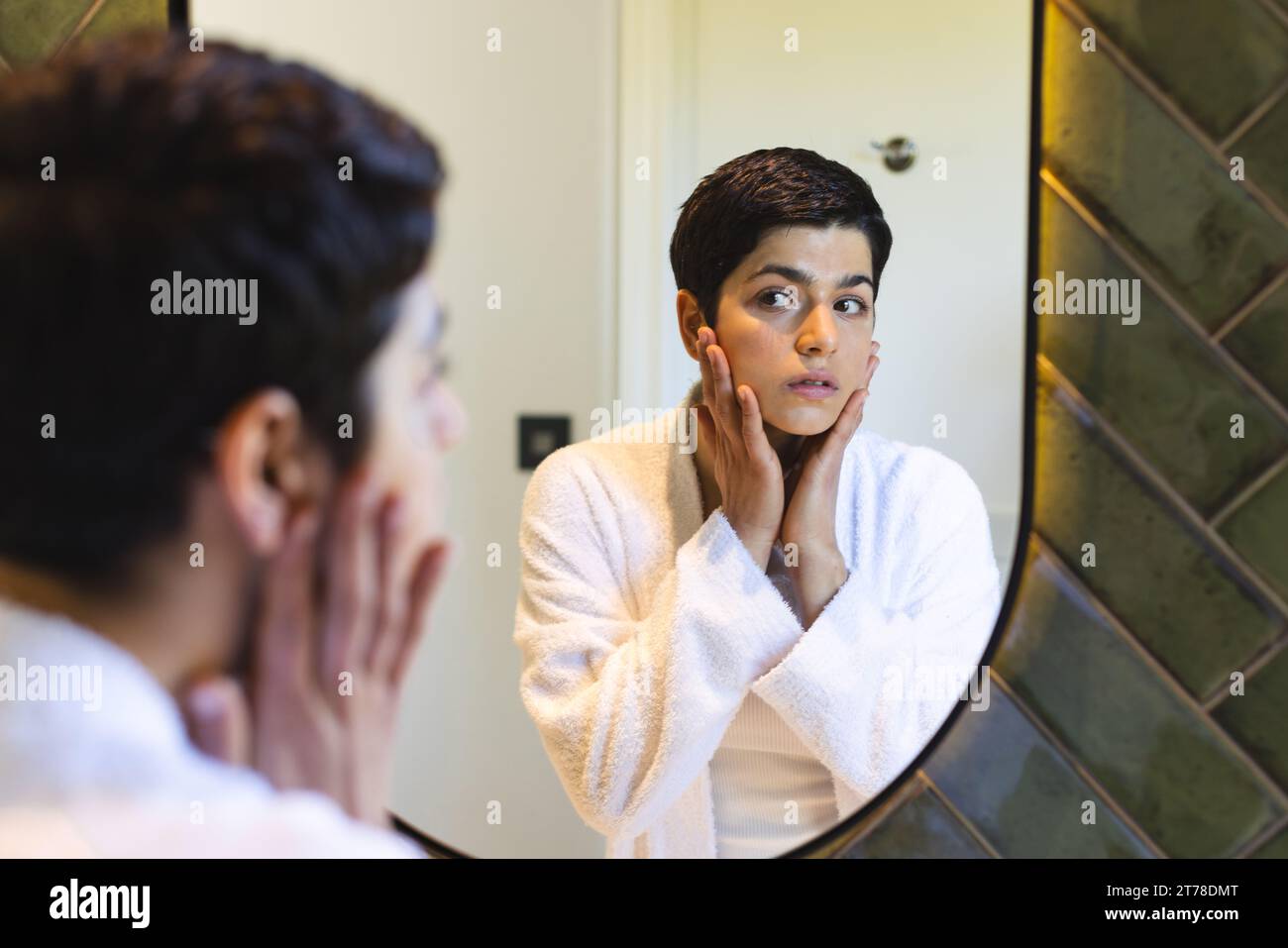 Glückliche birassische Frau, die das Gesicht berührt und zu Hause auf die Reflexion im Badezimmerspiegel blickt Stockfoto