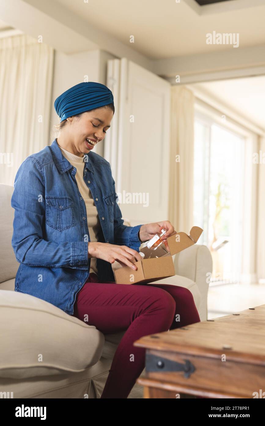 Glückliche birassische Frau im Hijab-Auspacken mit Kosmetikflasche im Wohnzimmer Stockfoto