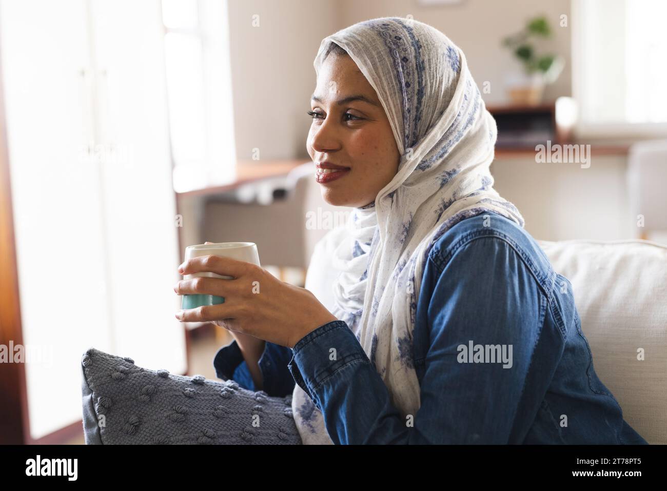 Glückliche birassische Frau im Hijab, die Kaffee im Wohnzimmer trinkt Stockfoto