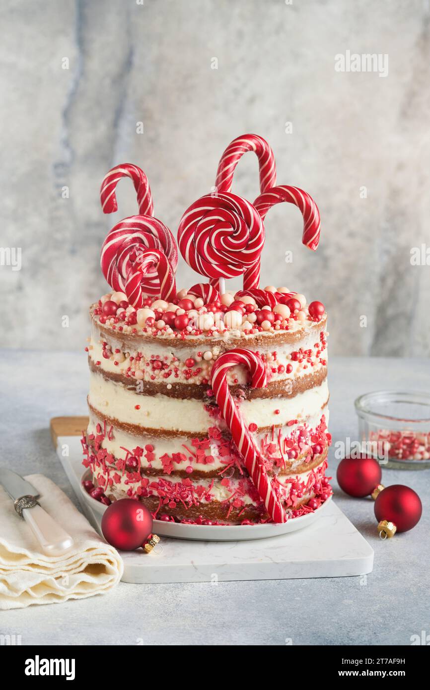 Mehrschichtiger Lebkuchenkuchen mit roten Lolli-Pops, farbigem Konfetti auf weißem Kuchenständer, Weihnachtskugeln, Tannenzweigen auf weißem Beton Stockfoto
