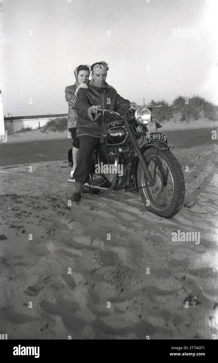 1950er Jahre, historisch, draußen, an einem Sandstrand, ein Mann in einer ledernen Bomberjacke, Armeetyp und Gesichtsbrille und eine Frau, die einen Schal trägt, sitzt zusammen auf einem Triumph Motorrad der Ära, England, Großbritannien. Das britische Motorrad wurde in Coventry von der Triumph Engineering Co hergestellt, deren Geschichte bis ins Jahr 1883 zurückreicht, und Siegfried Bettman, ein Immigrant aus Deutschland, der ein Import-/Exportgeschäft für Fahrräder gründete. Stockfoto