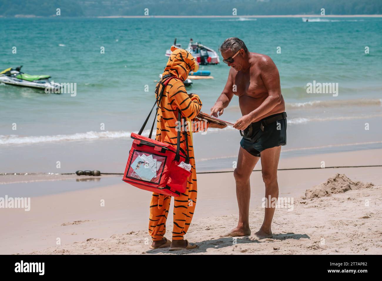 Verkäufer verkleidet als Orangentiger, der Eis an Touristen am Strand verkauft. Er spricht mit einem älteren Mann. PHUKET, THAILAND - 07. APRIL 2023 Stockfoto