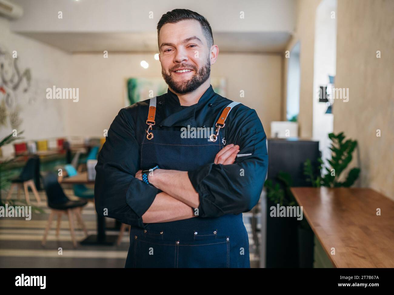 Porträt eines glücklichen, lächelnden kleinen Geschäftsinhabers in schwarzer Küchenuniform mit Schürze in seinem gemütlichen Restaurantsaal. Erfolgreiche Menschen, harte Arbeit Stockfoto