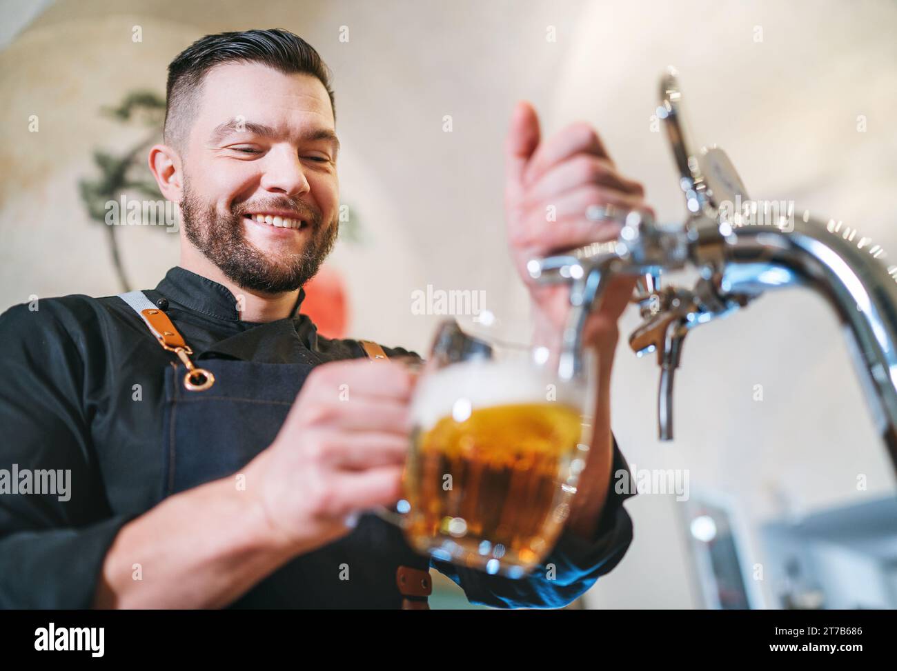 Ein lächelnder, stilvoller Bärenbarmann in schwarzer Uniform mit einer Schürze, der frisches Lagerbier in einen Pilsner-Glas-Becher an der Bar zapft. Erfolgreiche Mitarbeiter, Stockfoto